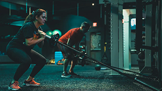 A woman performs a landmine squat in a dimly lit gym, holding a barbell anchored to the floor while a man exercises in the background near weight racks.
